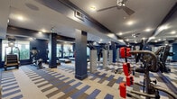 This fitness center in Panama City Beach, Florida, offers a well-appointed and comprehensive workout environment designed to support a wide range of exercise routines. The space is defined by bold navy blue walls, structural columns, and a multi-toned striped carpet in blue, gray, and beige that adds visual energy to the layout while delineating different workout zones throughout the floor.

The main exercise area is generously proportioned, with tray ceilings fitted with recessed lighting and ceiling fans that maintain air circulation throughout. Large windows along the perimeter walls allow natural light to filter in, offering views of the surrounding landscaped grounds and palm trees — a nod to the coastal Florida setting.

Full-length mirrors line several walls, enhancing the sense of space and providing functional feedback during strength training. The strength zone is stocked with a robust selection of cable machines, weight benches, resistance equipment, and free weights. A dedicated cardio section houses multiple treadmills, ellipticals, stair climbers, and stationary bikes equipped with digital displays.

A separate enclosed area within the facility provides additional space for focused workouts, complete with a barbell rack and supplementary machines. The overall ambiance is modern, functional, and energizing.