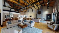 This expansive single-floor communal space masterfully blends industrial heritage with modern comfort. Soaring ceilings reveal an open truss structure with exposed wooden rafters and black steel ductwork, lending a dramatic loft-like atmosphere throughout. Original wide-plank hardwood floors run continuously across the entire space, anchoring the warm, inviting aesthetic.

The main lounge area is generously appointed with plush, neutral-toned sofas and armchairs arranged around patterned area rugs and a wooden coffee table — complete with a chess set — creating a relaxed social hub. A sleek linear gas fireplace anchors one wall, while a mounted television and a large decorative clock add character to the white-painted brick surround.

Raw exposed brick walls define key architectural moments throughout, including the entrance zone, which features floor-to-ceiling glass doors opening directly onto a furnished outdoor patio. A dedicated café corner, branded as a Starbucks station, is complemented by a full service bar with ample counter space, a sink, refrigeration, and beverage equipment. A separate business center offers workstations with computers and printing amenities in a windowed room with excellent natural light. A billiards table rounds out the recreational offerings. The space's abundant glazing and patio access ensure a seamless connection to the outdoors.