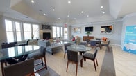 This single-floor space is thoughtfully designed as a multifunctional lounge and social area, combining comfort with a refined aesthetic. The room features a vaulted ceiling with recessed lighting that evenly illuminates the space, creating a bright and welcoming ambiance throughout.

Large windows along the perimeter allow generous amounts of natural light to fill the room, enhancing the airy, open feel. A classic fireplace serves as a focal point on one wall, flanked by the windows and complemented by decorative accents, adding warmth and character to the space.

The layout is organized into distinct seating zones. A lounge area features plush gray sofas, tufted ottomans, and accent chairs arranged around a glass coffee table, accented with orange throw pillows for a pop of color. Nearby, round glass-topped dining tables paired with upholstered chairs provide casual seating suitable for dining or conversation.

A wall-mounted flat-screen television is positioned above a wooden credenza, adding a media-friendly element to the space. Contemporary artwork adorns the walls, contributing to the polished, modern character. A patterned area rug anchors the central seating arrangement, tying the design elements together cohesively.