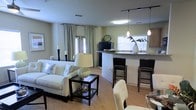 This single-floor residence showcases an open-concept layout that integrates the living, dining, and kitchen areas into one cohesive, flowing space. The living area is anchored by a plush white loveseat and complemented by a sleek black coffee table, creating a clean, contemporary aesthetic. Large windows dressed with striped curtains invite abundant natural light, lending the space a bright and airy atmosphere. Double doors at the far end of the living area provide additional light and potential outdoor access.

The kitchen features a curved breakfast bar with dark granite countertops, accommodating tall bar-height seating for casual dining. Pendant lighting hangs above the bar, adding focused illumination and a stylish touch. The kitchen is also equipped with track lighting overhead, cabinetry in warm tones, and a window above the sink that draws in further natural light.

Light hardwood-style flooring runs throughout the space, unifying each zone visually while enhancing the overall warmth of the interior. The neutral color palette of soft greens and whites, combined with carefully chosen décor accents, gives this residence a polished, welcoming ambiance well-suited for everyday living and entertaining.