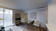 This single-floor space presents a clean, minimalist design centered around an open-plan layout that combines living and dining functions. The room features smooth, light-painted walls and a flat white ceiling that contribute to a bright, airy atmosphere, complemented by natural light filtering in through a sliding glass door or large window along one wall.

The dining area is anchored by a glass-topped table with a sleek metal frame, surrounded by a mix of seating including an adjustable office-style chair and tan upholstered dining chairs, offering a versatile setup suited for both dining and work. A framed botanical line-art print adorns the wall, adding a subtle decorative touch.

A black metal open shelving unit along the adjacent wall provides storage and display space, currently holding a small appliance and decorative items. A wall-mounted split-system air conditioning unit ensures year-round climate control. The flooring throughout is wood-look planking in a warm, medium tone, adding warmth and continuity to the space. The overall aesthetic is modern and understated, with a functional, well-organized feel.