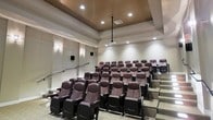 This dedicated screening room delivers a true theater experience within a private setting. The space is designed with tiered, stadium-style seating arranged in ascending rows, ensuring unobstructed sightlines from every position. The plush, dark burgundy upholstered seats feature wide armrests with integrated cupholders, prioritizing comfort during extended viewing sessions.

A ceiling-mounted projector is centrally positioned to project onto the front screen wall, supported by recessed lighting that provides ambient illumination throughout the room. The ceiling incorporates a distinctive perforated acoustic panel inset, which aids in sound absorption and overall audio performance. Wall-mounted speakers on either side of the room further enhance the sound system, contributing to a well-balanced acoustic environment.

The walls are finished in a neutral, light tone that complements the darker seating, while carpeted stepped aisles provide safe, easy access between rows. Subtle wall sconces add a classic cinematic ambiance. Overall, the room is thoughtfully designed to replicate the feel of a professional movie theater while maintaining an intimate, upscale atmosphere.