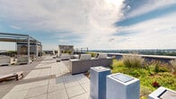Located atop 42 Broad St W in Mount Vernon, NY, this thoughtfully designed rooftop terrace offers an exceptional outdoor experience with a variety of distinct zones for relaxation and entertaining. The space is defined by its lush, integrated landscaping — native ornamental grasses and plantings line the perimeter walls, creating a green, garden-like atmosphere high above the cityscape.

A generous artificial turf lawn area provides a comfortable setting for casual lounging, furnished with wooden deck chairs for residents to enjoy the open sky and fresh air. Adjacent to this, a hardwood deck hosts an elegant outdoor living room anchored by a striking stacked-stone accent wall with a built-in fire feature. Plush white modular seating surrounds the space, while a pergola structure overhead adds architectural definition.

The terrace also features a well-equipped outdoor kitchen and grill station, ideal for al fresco dining. Throughout the space, panoramic views extend across the surrounding neighborhoods, treetops, and the broader New York metropolitan skyline, offering an impressive visual backdrop from every vantage point. The combination of sophisticated design, abundant greenery, and unobstructed views makes this rooftop terrace a standout amenity.