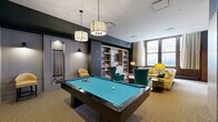 Located at 317 North Broad Street in Philadelphia, this single-floor communal space combines a well-appointed kitchen with a multifunctional living and entertainment area. The open-concept layout makes efficient use of its footprint, creating a welcoming environment ideal for socializing and recreation.

The living area is anchored by a full-size pool table with a teal felt surface, illuminated by stylish pendant fixtures overhead. A dedicated cue rack is built into the wall nearby for easy access. Opposite the game area, a comfortable lounge zone features a large sectional sofa, teal wingback chairs, and bold yellow accent seating, all arranged around low-profile coffee tables. Built-in bookshelves line the walls alongside a wall-mounted flat-screen TV, while large windows dressed with floor-length curtains flood the space with natural light and offer views of the surrounding streetscape.

The kitchen is defined by its clean, modern aesthetic, featuring a spacious central island with a white countertop and teal bar stools. Stainless steel appliances, including a side-by-side refrigerator, cooktop, and wall oven, complement the open shelving with under-cabinet lighting. Warm wood-tone wall paneling and stone-look tile flooring add texture and depth, tying the two zones together into a cohesive, inviting communal retreat.