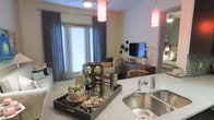 This single-floor residence is designed around an open-concept floor plan that seamlessly connects the kitchen and living areas. The kitchen features granite or stone-finish countertops and a double-basin stainless steel sink, providing a functional and polished workspace. A decorative bar tray with a wine rack, glassware, and cocktail tools adds a sophisticated entertaining touch to the counter space.

Overhead, vibrant red pendant lights hang from the ceiling, creating warm focal points that contrast with the neutral wall tones throughout the space. A ceiling fan is visible in the living area, contributing to both comfort and airflow.

The living area is furnished with a comfortable sofa and coordinating accent pieces, while a wall-mounted flat-screen television sits atop a media console accompanied by a desk and chair. Large windows dressed in layered curtains and blinds allow generous natural light to fill the space, creating a bright and welcoming ambiance. The overall design balances practicality with a modern, curated aesthetic that makes the space feel both livable and thoughtfully styled.