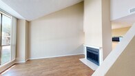 This well-designed two-story residence offers one bedroom and two full bathrooms across a thoughtfully arranged floor plan. The main level features an open living area distinguished by soaring vaulted ceilings that create an impressive sense of vertical space. A classic fireplace with a white mantle and tile surround serves as the focal point of the living room, which is finished with warm hardwood-style flooring throughout. A large sliding glass door floods the space with abundant natural light while providing direct views of mature trees and lush greenery outside. The kitchen is efficiently laid out with white cabinetry, light countertops, a double sink, and black appliances including a range, microwave, and refrigerator. A peninsula counter connects the kitchen to a dining or hallway area, enhancing the open flow of the main level. A full bathroom on this floor features a tub/shower combination with white tile surround. Upstairs, the generously sized primary bedroom features plush carpeting, a built-in desk area, and direct access to a well-appointed en suite bathroom with a wide vanity, ample cabinetry, and a lighted mirror. The upper level also includes a dedicated closet. Throughout the home, neutral warm tones and textured ceilings contribute to a clean, move-in-ready ambiance.