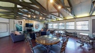 This single-floor community space makes an immediate impression with its striking vaulted ceiling, where bold exposed structural trusses painted in deep green and wood tones span the length of the room. Track lighting mounted along the beams illuminates the open interior, complementing the natural light that pours in through a generous row of windows along one wall, offering views of surrounding trees and greenery.

The main hall is defined by rich, polished concrete floors in a deep reddish-brown hue, creating a warm and durable foundation throughout. A prominent stone fireplace anchors the lounge area, which features comfortable upholstered sofas and armchairs arranged around a wooden coffee table. Large-format artwork depicting vintage street scenes adds a curated, inviting character to the walls.

The space transitions seamlessly into a flexible dining and gathering area, furnished with multiple round and rectangular tables paired with a variety of seating options, accommodating both intimate gatherings and larger group events. A dedicated kitchen area supports catering and event use, equipped with stainless steel appliances including a refrigerator, range, and microwave, along with a central island and ample cabinetry. The overall ambiance is open, versatile, and well-suited for community gatherings, meetings, or social events.