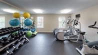Located at 553 S Commercial in Manchester, New Hampshire, this well-equipped fitness center offers a comprehensive single-floor facility designed to support a wide range of workout routines. The space features durable speckled rubber flooring throughout, providing both safety and comfort during exercise. Overhead flush-mounted lighting panels illuminate the entire area evenly, creating a bright and functional environment.

The cardio zone is stocked with multiple treadmills, upright and recumbent stationary bikes, and elliptical machines, many of which are equipped with individual monitor screens for entertainment during workouts. A multi-station cable weight machine anchors the strength training area, accompanied by a free weight rack with a graduated dumbbell selection and weight benches for a full resistance training experience. A large wall-mounted mirror reflects the strength zone, enhancing the sense of space and allowing users to monitor their form.

A dedicated accessories area features a colorful rack of stability and medicine balls in varying sizes, resistance bands, and foam rollers, catering to flexibility, balance, and recovery needs. Natural light filters in through windows on one wall, adding to the open and welcoming atmosphere. A separate entry door with emergency exit signage ensures safe and convenient access throughout the facility.