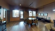 Located at 4701 Seventh Ave SW in Olympia, Washington, this single-floor community amenity space is thoughtfully designed for recreation and social gathering. The layout flows seamlessly across several interconnected areas, all unified by warm earth-tone walls and rich hardwood-style flooring that lend a welcoming, relaxed ambiance throughout.

The centerpiece of the space is a well-appointed game room featuring a full-size pool table illuminated by a striking pendant light fixture, alongside a shuffleboard table. A gallery of framed billiard-themed artwork lines the walls, adding a polished, curated touch. Natural light filters in through large windows, offering views of the surrounding neighborhood.

Adjacent to the game room is a casual lounge and dining area furnished with a mix of round and rectangular tables, upholstered chairs, and comfortable booth-style seating. Track lighting and pendant fixtures create a warm, layered glow, while additional windows enhance the bright and open feel. A flat-screen television mounted above a distressed wood console provides entertainment options in the lounge corridor.

A separate card and game table area, surrounded by plush seating and bathed in natural light from multiple windows, offers an ideal setting for group activities. The space also includes access to an exterior door, suggesting patio or outdoor connectivity. Overall, this amenity space delivers a versatile, well-equipped environment for leisure and community engagement.
