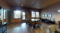 Located at 4701 Seventh Ave SW in Olympia, Washington, this single-floor community amenity space is thoughtfully designed for recreation and social gathering. The layout flows seamlessly across several interconnected areas, all unified by warm earth-tone walls and rich hardwood-style flooring that lend a welcoming, relaxed ambiance throughout.

The centerpiece of the space is a well-appointed game room featuring a full-size pool table illuminated by a striking pendant light fixture, alongside a shuffleboard table. A gallery of framed billiard-themed artwork lines the walls, adding a polished, curated touch. Natural light filters in through large windows, offering views of the surrounding neighborhood.

Adjacent to the game room is a casual lounge and dining area furnished with a mix of round and rectangular tables, upholstered chairs, and comfortable booth-style seating. Track lighting and pendant fixtures create a warm, layered glow, while additional windows enhance the bright and open feel. A flat-screen television mounted above a distressed wood console provides entertainment options in the lounge corridor.

A separate card and game table area, surrounded by plush seating and bathed in natural light from multiple windows, offers an ideal setting for group activities. The space also includes access to an exterior door, suggesting patio or outdoor connectivity. Overall, this amenity space delivers a versatile, well-equipped environment for leisure and community engagement.