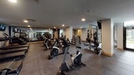 Located at 240 Park Ave in Minneapolis, Minnesota, this fitness center is a fully outfitted, single-floor facility designed to support a wide range of workout routines. The space is organized into distinct functional zones, each dedicated to a specific training discipline.

The cardio area features a row of treadmills, elliptical trainers, and stationary bikes, all equipped with individual monitors mounted on the walls above, allowing users to stay entertained during their sessions. Recessed ceiling lighting illuminates the space throughout, creating a consistent and focused workout environment.

The free weights section includes a comprehensive dumbbell rack with a full range of weights, accompanied by a flat workout bench for versatile strength training. Full-length mirrors line the walls in this area, enhancing the sense of space and allowing users to monitor their form.

The strength and functional training zone houses a variety of cable machines, a power tower for bodyweight exercises, and plate-loaded equipment, providing ample options for resistance training. Structural columns are integrated throughout the floor plan, and the layout ensures clear sightlines and comfortable circulation between equipment.

Rubber tile flooring covers the entire facility, offering both durability and safety underfoot. Large windows along one side of the space allow natural light to filter in, adding an open, airy quality to the otherwise sleek, dark-toned interior.