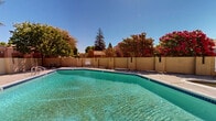 Located at 1540 Mosaic Way in Stockton, California, this outdoor community pool area offers a well-maintained recreational space designed for relaxation and leisure. The centerpiece is a generously sized swimming pool with clear, blue-green water, surrounded by a broad concrete deck that provides ample room for poolside activities. The pool area is enclosed by a tan stucco perimeter wall that ensures privacy while complementing the warm architectural tones of the adjacent multi-story residential buildings.

Access to the pool is secured through a gated entry featuring metal railings and a staircase, adding both functionality and a sense of structure to the space. The surrounding landscape is enriched by mature trees — including tall pines and vibrant flowering bougainvillea — that frame the area with lush greenery and seasonal color, creating a pleasant natural backdrop. The warm California sunshine floods the open space, making it an inviting outdoor retreat throughout the year.

The pool area also includes modest amenities such as seating and utility areas positioned along the perimeter. The overall ambiance is one of casual comfort, with the combination of clear water, open skies, mature vegetation, and Mediterranean-inspired stucco architecture evoking a relaxed, resort-like atmosphere suited to the sunny Stockton climate.