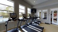 Located at 720 Michigan Ave in Columbus, Ohio, this fitness center offers a well-equipped and welcoming environment for residents looking to maintain an active lifestyle. The space is outfitted with a row of True-brand treadmills and additional cardio machines, providing ample options for cardiovascular training. Large windows line the walls, flooding the room with natural light and offering views of the outdoor pool and surrounding grounds, creating an open and energizing atmosphere. A wall-mounted flat-screen television is positioned for easy viewing during workouts, adding to the comfort and convenience of the facility. The interior features neutral gray walls, recessed lighting, and patterned carpeted flooring that lend the space a clean, polished appearance. A large mirror on one wall enhances the sense of openness and allows users to monitor their form. A glimpse through the mirror reveals an adjacent area with additional amenities, suggesting a broader community facility. The overall ambiance is bright, functional, and thoughtfully designed to support a complete fitness experience.