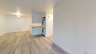 This single-floor residence offers a clean, modern aesthetic throughout, combining practical design with comfortable living. The main living area features light-toned wood-look flooring and crisp white walls, creating a bright and open atmosphere. A baseboard heating system runs along the living space wall, providing efficient warmth. The kitchen is thoughtfully designed with white shaker-style cabinetry, gray countertops, a stainless steel sink, dishwasher, and range. A window above the sink allows natural light to filter in. Conveniently situated adjacent to the kitchen is an in-unit laundry area with a stacked washer and dryer tucked neatly into a dedicated alcove. The bedroom is carpeted for added comfort and features a ceiling fan with a light fixture, a large window offering views of surrounding greenery, and a mirrored closet. The bathroom is finished in clean white tones with wood-look flooring, featuring a full tub and shower combination with chrome fixtures and a towel bar. The overall layout, as seen in the dollhouse view, is compact and efficient, making excellent use of the available space while maintaining a fresh, move-in-ready condition throughout.