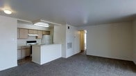 This single-floor space offers a practical and functional open-concept layout designed to maximize usability. The kitchen is positioned along one side of the main living area and features light wood cabinetry, a white refrigerator, a gas range, and a built-in overhead fluorescent light fixture that brightens the workspace. A white peninsula counter separates the kitchen from the living area, providing a natural division between cooking and living spaces while maintaining an open, connected feel. The living area is generously sized, finished with neutral wall-to-wall carpeting that adds warmth and comfort underfoot. A large HVAC unit is mounted flush against the wall, ensuring year-round climate control. Crisp white walls throughout give the space a clean, bright aesthetic. A hallway visible from the living area leads toward additional rooms, which appear to benefit from natural light filtering in through a window at the far end. The overall layout is straightforward and efficient, making it well-suited for everyday living. The neutral color palette and simple finishes provide a versatile backdrop that can be easily personalized.