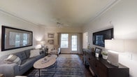 This single-floor residence offers a thoughtfully designed interior that balances comfort and style. The living room serves as the heart of the space, showcasing rich hardwood flooring, crisp crown molding, and a ceiling fan that adds both function and a classic architectural touch. A pair of French doors at the far end of the room flood the space with natural light while providing a welcoming view of the outdoors, framed by elegant patterned drapes. The living area is generously proportioned, comfortably accommodating a full seating arrangement centered around an oval coffee table atop a layered area rug. Soft, neutral wall tones complement the warm wood tones throughout, creating a cohesive and inviting ambiance. A substantial dark-wood credenza along one wall provides ample storage and display space, topped with decorative accents and anchored by a large wall-mounted television. Tasteful framed artwork adorns the walls, contributing to the refined yet approachable character of the room. Warm accent lighting from table lamps enhances the cozy atmosphere, making the space ideal for both relaxed everyday living and entertaining guests.