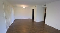 This single-story space offers a clean, open layout defined by rich, dark hardwood flooring that extends throughout the main living area, creating a warm contrast against the bright white walls and ceiling. A centrally mounted ceiling light fixture provides general illumination, contributing to the well-lit, airy feel of the room. The walls are freshly finished in a neutral white, offering a versatile backdrop suitable for a variety of interior styles.

The open floor plan allows for a natural flow from the main living area toward adjacent spaces, including what appears to be a hallway leading to additional rooms and a partially visible area to the right that hints at a kitchen or utility space. Architectural details such as visible ceiling vents indicate a central HVAC system for year-round climate control.

The layout is straightforward and functional, with clean lines and minimal visual clutter that emphasize the sense of openness. White baseboards frame the flooring neatly throughout, adding a polished finishing touch. Overall, the space presents a move-in-ready environment with a neutral palette and durable finishes well-suited for everyday living.