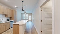 Located at 14000 Tapestry Dr in Orlando, Florida, this one-bedroom, one-bathroom residence offers a thoughtfully designed single-floor layout with a clean, modern aesthetic throughout. The open-concept kitchen and living area serves as the heart of the home, featuring light wood-tone cabinetry, stainless steel appliances including a refrigerator, range, and microwave, a white subway tile backsplash, and a central island with a bright white countertop. Sleek pendant lighting and recessed fixtures illuminate the space, while light hardwood-style flooring extends across the main living areas. The living room benefits from natural light through large windows with blinds and direct access to an outdoor balcony. The bedroom is a comfortable retreat with soft wall-to-wall carpeting, a ceiling fan, and a window offering outdoor views of surrounding greenery. The bathroom is well-appointed with light wood vanity cabinetry, a white countertop, a glass-enclosed walk-in shower with white subway tile, built-in shelving, and matching hardwood-style flooring. A dedicated in-unit laundry room houses a full-size top-loading washer and dryer with overhead wire shelving for added storage. A separate closet rounds out the functional layout of this well-finished Orlando residence.