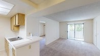 This single-floor unit offers a well-organized, open layout that connects the main living and kitchen areas seamlessly. The kitchen is a functional galley-style space equipped with light oak cabinetry, granite-patterned countertops, a gas range, a full-size refrigerator, a dishwasher, and a sink integrated into a peninsula that faces the living area. A recessed skylight panel above the kitchen provides bright, even overhead illumination, enhancing the sense of openness. The kitchen floor is finished with neutral tile, providing a clean transition to the carpeted living and bedroom areas. The living room is generously proportioned, featuring soft wall-to-wall carpeting, a ceiling fan, and multiple large windows dressed with vertical blinds that allow ample natural light to fill the space. The front entry is positioned within the living area and is flanked by additional windows, contributing to the airy, welcoming atmosphere. The bedroom is a comfortable, carpet-floored room with its own window and vertical blinds, offering a quiet and private retreat. The bathroom includes a full tub-and-shower combination with tile surround, a toilet, a granite-patterned countertop vanity, and a large mirror. Throughout, the unit maintains a clean, neutral palette of whites and beiges, creating a bright and move-in-ready environment.