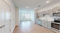 This single-floor residence presents a clean, contemporary design characterized by a bright, open layout and a cohesive neutral palette throughout. The open-concept living and kitchen area serves as the heart of the space, where light wood-style flooring extends seamlessly underfoot, complemented by white walls and recessed lighting. A ceiling fan anchors the living area, which receives abundant natural light through large windows offering views of the surrounding community.

The kitchen is well-appointed with gray shaker-style cabinetry, white subway tile backsplash, light countertops, and a full suite of stainless steel appliances including a refrigerator with ice maker, electric range, microwave, and dishwasher. A double-basin sink with a modern faucet completes the functional workspace.

The bedroom features plush wall-to-wall carpeting, a ceiling fan, and windows that bring in natural light. An en-suite bathroom offers a gray vanity with a large framed mirror, white countertop, wood-style flooring, and a full soaking tub with subway tile surround. A closet with double doors provides generous storage near the entryway, which opens into the main living space.