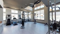 The Cardio Center is a thoughtfully designed fitness facility that combines functionality with an open, energizing atmosphere. The space features soaring vaulted ceilings accentuated by large clerestory windows along the upper walls, which flood the interior with natural light and create an airy, expansive feel. Oversized windows along the main walls offer views of the surrounding outdoor environment, keeping the space bright and connected to the exterior.

The light-toned hardwood-style flooring contributes to the clean, modern aesthetic throughout the center. Ceiling fans are strategically positioned to maintain comfortable airflow during workouts. The facility is well-appointed with a comprehensive selection of cardio and strength-training equipment, including treadmills, elliptical machines, and cable weight systems, providing users with a full range of workout options. The equipment is arranged to allow ample space for movement and circulation between stations.

The neutral color palette of warm whites and soft grays, combined with the generous natural light, creates an inviting and motivating environment. The overall design prioritizes both practicality and comfort, making the Cardio Center a versatile and accessible fitness space suited for a wide variety of exercise routines.