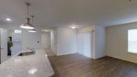 This single-floor space showcases a clean, modern design with an open-concept layout that connects the kitchen and living areas seamlessly. The kitchen features a granite countertop with a stainless steel sink and stylish pendant lighting overhead, adding a contemporary touch. The living area is finished with warm wood-style flooring that runs throughout, complementing the bright white walls and white baseboards. A sliding door fitted with vertical blinds allows for natural light control and likely provides access to an outdoor area. Flush-mount ceiling lighting keeps the space well-lit, while a hallway visible in the background leads to additional rooms, suggesting a practical and functional floor plan. The neutral color palette and clean lines throughout give the space a fresh, move-in-ready feel. Overall, the open layout and modern finishes create a welcoming and versatile environment suited for everyday living.