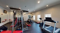Located at 1816 Wards Ferry Rd in Lynchburg, Virginia, this fitness center is a comprehensively outfitted, single-floor facility designed to support a wide range of workout routines. The space is finished with durable rubber flooring throughout, providing both safety and stability for exercise. Recessed lighting illuminates the room evenly, while multiple windows along the walls allow natural light to filter in and offer views of the surrounding exterior.

The main workout area is generously proportioned, housing a multi-station cable machine with red-accented benches and attachments suitable for a full range of strength training exercises. Cardio equipment is well-represented, with treadmills, elliptical trainers, and a stationary bike positioned across the floor. A dedicated free weights section includes a tiered dumbbell rack stocked with a variety of weights, accompanied by a flat bench and a preacher curl station. A large wall mirror spans one side of the room, enhancing the sense of space and allowing users to monitor their form during workouts. A wall-mounted flat-screen television adds convenience for entertainment during exercise. The clean, white walls and ceiling contribute to a bright, motivating atmosphere throughout the facility.