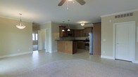 This single-story home showcases an open-concept design that seamlessly connects the living area and kitchen, creating a bright and functional space. The kitchen is a standout feature, outfitted with rich dark wood cabinetry that extends to the upper walls, dark granite countertops, and a full suite of stainless steel appliances, including a gas range with hood, built-in microwave, and a full-size refrigerator. A central island with matching cabinetry provides additional prep space and serves as a natural divider between the kitchen and living areas. Pendant lighting hangs above the island, complementing the flush-mount fixture nearby. The living area is finished with neutral carpet and features a ceiling fan, adding to the room's comfort. Crown molding runs throughout, lending a polished, cohesive look to the interior. Large tile flooring covers the kitchen and surrounding areas, offering durability and a clean aesthetic. A doorway visible from the main living space leads to additional rooms, and natural light filters in from windows, contributing to the home's open and airy ambiance.