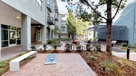This expansive outdoor courtyard is thoughtfully designed to serve as a central gathering and recreation hub. The space is defined by warm brick-paver pathways that wind through carefully landscaped garden beds featuring mature trees, ornamental grasses, and layered shrubs, creating a lush, park-like atmosphere. Multiple distinct zones cater to varied activities and social settings. A dedicated grilling station anchors one end of the courtyard, equipped with built-in gas grills set beneath a striking laser-cut metal pergola with an intricate geometric canopy, paired with a sleek wood-slatted communal bar table and stools for outdoor dining. Nearby, a decorative water feature framed by mosaic tile serves as a visual focal point, surrounded by a dining set ideal for al fresco meals. Casual seating vignettes are distributed throughout, featuring modern woven lounge chairs and cylindrical side tables, providing intimate spaces for relaxation. A dedicated games area includes oversized lawn games such as cornhole and a stacking block tower. Signage throughout indicates additional amenities including a games room and bike storage, accessible directly from the courtyard. The surrounding multi-story building with arched entryways and balconies encloses the space, creating a sheltered, community-oriented environment.