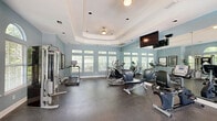 Located at 6650 Cottage Hill Rd in Mobile, Alabama, this fitness center offers a thoughtfully designed exercise space for residents seeking a convenient on-site workout facility. The room is defined by a distinctive tray ceiling with recessed lighting and a ceiling fan, lending an architectural elegance uncommon in typical gym settings. Soft blue-gray walls create a calm, motivating atmosphere throughout the space.

Natural light floods the room through a series of large windows positioned along multiple walls, offering pleasant outdoor views of surrounding greenery. Glass-paneled doors provide direct access to the exterior, enhancing both ventilation and connectivity to outdoor areas. Full-length mirrors along one wall amplify the sense of space and allow users to monitor their form during workouts.

The facility is equipped with a comprehensive selection of cardio machines, including treadmills, ellipticals, and a recumbent bike, primarily from the True Fitness brand. A multi-station cable strength-training machine rounds out the equipment offerings for a full-body workout experience. A wall-mounted flat-screen television adds entertainment options for users during exercise sessions. Durable rubber flooring throughout provides comfort and safety underfoot. Framed artwork on the walls adds a refined finishing touch to this well-appointed fitness center.