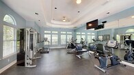 Located at 6650 Cottage Hill Rd in Mobile, Alabama, this fitness center offers a thoughtfully designed exercise space for residents seeking a convenient on-site workout facility. The room is defined by a distinctive tray ceiling with recessed lighting and a ceiling fan, lending an architectural elegance uncommon in typical gym settings. Soft blue-gray walls create a calm, motivating atmosphere throughout the space.

Natural light floods the room through a series of large windows positioned along multiple walls, offering pleasant outdoor views of surrounding greenery. Glass-paneled doors provide direct access to the exterior, enhancing both ventilation and connectivity to outdoor areas. Full-length mirrors along one wall amplify the sense of space and allow users to monitor their form during workouts.

The facility is equipped with a comprehensive selection of cardio machines, including treadmills, ellipticals, and a recumbent bike, primarily from the True Fitness brand. A multi-station cable strength-training machine rounds out the equipment offerings for a full-body workout experience. A wall-mounted flat-screen television adds entertainment options for users during exercise sessions. Durable rubber flooring throughout provides comfort and safety underfoot. Framed artwork on the walls adds a refined finishing touch to this well-appointed fitness center.