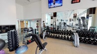 This contemporary fitness center is designed to meet the needs of a wide range of fitness enthusiasts, offering a comprehensive suite of equipment within a bright, open layout. The space features soaring ceilings that lend an airy, expansive feel, complemented by large windows that usher in abundant natural light throughout.

The free weights area is generously appointed, housing multiple racks of dumbbells spanning a full range of weights, kettlebells, medicine balls, and a BOSU balance trainer. Adjustable benches and a squat/barbell rack provide dedicated space for strength training, while floor-to-ceiling mirrors along the walls enhance the sense of space and allow for proper form monitoring.

A dedicated cardio zone lines one side of the facility, featuring a row of treadmills and other aerobic machines. A multi-functional cable/pulley training station anchors the center of the floor, offering versatile options for resistance and functional training. Ceiling fans and recessed lighting ensure a comfortable workout environment at all times.

Mounted flat-screen televisions are positioned throughout the space for entertainment, and a digital timer display keeps users on track during their sessions. The durable, speckled rubber-composite flooring provides safety and comfort underfoot. The overall aesthetic is clean, modern, and professional, creating an inviting atmosphere for daily training.