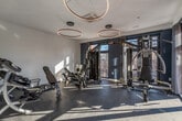 This contemporary fitness center is thoughtfully designed to deliver a high-end workout experience. The space is anchored by a striking set of circular ring pendant lights suspended from white ceilings, providing both functional illumination and a bold architectural focal point. The floors are finished with durable interlocking rubber tiles, offering both safety and practicality throughout the workout area.

The gym is well-appointed with a variety of professional-grade strength equipment, including a multi-functional cable machine, adjustable weight benches, a leg press machine, and a dumbbell rack, catering to a full range of strength and conditioning exercises. A dedicated cable and pulley system is positioned along one side, maximizing the versatility of the training space.

Large floor-to-ceiling windows and glass-panel doors line one wall, inviting abundant natural light and offering views of the surrounding outdoor environment. The accent wall is finished in a deep navy blue, adding a modern contrast to the bright white walls and ceiling. Entry to the fitness center is through sleek, full-glass doors with dark metal frames, reinforcing the space's contemporary aesthetic. The overall ambiance is clean, bright, and purpose-built for focused, effective training.