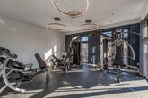 This contemporary fitness center is thoughtfully designed to deliver a high-end workout experience. The space is anchored by a striking set of circular ring pendant lights suspended from white ceilings, providing both functional illumination and a bold architectural focal point. The floors are finished with durable interlocking rubber tiles, offering both safety and practicality throughout the workout area.

The gym is well-appointed with a variety of professional-grade strength equipment, including a multi-functional cable machine, adjustable weight benches, a leg press machine, and a dumbbell rack, catering to a full range of strength and conditioning exercises. A dedicated cable and pulley system is positioned along one side, maximizing the versatility of the training space.

Large floor-to-ceiling windows and glass-panel doors line one wall, inviting abundant natural light and offering views of the surrounding outdoor environment. The accent wall is finished in a deep navy blue, adding a modern contrast to the bright white walls and ceiling. Entry to the fitness center is through sleek, full-glass doors with dark metal frames, reinforcing the space's contemporary aesthetic. The overall ambiance is clean, bright, and purpose-built for focused, effective training.