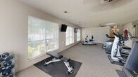 Located at 5100 S Zero St in Fort Smith, Arkansas, this dedicated exercise room offers a comfortable and functional environment for a well-rounded workout. The space is carpeted throughout and features a textured ceiling with ceiling fans that help maintain airflow during exercise sessions. Multiple large windows fitted with blinds line the walls, allowing generous natural light to fill the room while providing views of the surrounding greenery outdoors. Wall-mounted flat-screen televisions are positioned at convenient viewing angles, enhancing the workout experience. The fitness area is thoughtfully arranged with a variety of equipment, including treadmills, elliptical machines, a recumbent stationary bike, and a weight bench with a rubber mat, complemented by a dumbbell rack stocked with free weights. A mirrored wall on one side adds depth to the space and supports strength training. Decorative sports-themed artwork adorns the neutral-toned walls, giving the room a lively, motivating atmosphere. Built-in storage is provided by a set of white panel doors along one wall. The room is clearly signed and accessible, with an exit indicator ensuring safety. Overall, this exercise room delivers a practical and inviting setting for daily fitness routines.
