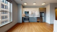 Located at 66 Fountain St in the heart of Providence, Rhode Island, this modern single-floor unit offers a clean, contemporary design throughout. The open living area is anchored by warm hardwood flooring and soft gray walls, creating a bright and welcoming atmosphere. A large window frames views of the surrounding urban skyline, filling the space with natural light.

The kitchen is a standout feature, thoughtfully designed with two-tone cabinetry — sleek white upper cabinets paired with muted blue-gray lower cabinets — and a white quartz countertop. A hexagonal tile backsplash adds texture and visual interest, while stainless steel appliances, including a refrigerator, range, microwave, and dishwasher, provide a fully equipped cooking space. Flush-mount ceiling lights illuminate the area evenly.

The layout flows naturally from the kitchen into the open living space, which benefits from a split-level design element that adds architectural dimension. The bathroom is finished with neutral large-format tile flooring and features a clean, minimalist aesthetic consistent with the rest of the unit. Throughout the space, modern flat-panel doors, recessed baseboards, and fire safety fixtures reflect a well-considered, up-to-date build. This unit offers a polished urban living environment in a prime Providence location.