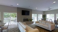 This single-floor space is designed with a clean, contemporary aesthetic that emphasizes comfort and openness. The main living area serves as the focal point of the space, anchored by a modern fireplace set within a floor-to-ceiling gray accent wall. A large flat-screen television is mounted prominently above the fireplace, creating a well-defined entertainment zone. The seating arrangement features cream-colored sofas positioned on a patterned area rug, contributing to the room's neutral, sophisticated color palette.

Recessed lighting in the smooth white ceiling provides even illumination throughout, complementing the abundant natural light that streams in through multiple large sliding glass doors along the perimeter. These doors offer views of an outdoor seating area surrounded by lush greenery, effectively blurring the boundary between indoor and outdoor living. Additional potted plants are placed near the fireplace, adding a touch of organic warmth to the otherwise minimalist interior. The overall ambiance is calm, airy, and well-appointed, making the space well-suited for both relaxation and casual gatherings.