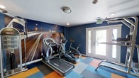 This fitness center is designed to energize and inspire, combining functional equipment with dynamic visual elements. The space is anchored by a striking full-wall mural depicting a dramatic urban highway scene at night, with streaks of light lending a sense of motion and vitality to the room. Deep navy blue walls complement the mural and create an immersive, high-energy atmosphere throughout.

The cardio area is well-appointed with a treadmill, elliptical trainer, and stationary bike, providing a well-rounded selection of aerobic equipment for users of varying fitness levels. Additional exercise equipment is visible, suggesting the space accommodates a range of workout routines.

Underfoot, a colorful patchwork of modular carpet tiles in shades of blue, orange, and gray adds a playful, modern touch to the flooring, enhancing the room's contemporary aesthetic. Overhead, flush-mounted lighting and an illuminated exit sign ensure the space is well-lit and safety-conscious.

A set of glass-paneled French doors along one wall floods the room with natural light and provides direct access to an outdoor area, offering ventilation and a refreshing connection to the outdoors. The single-floor layout makes the space easily accessible, delivering a clean, focused environment purpose-built for fitness.