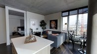 This contemporary single-floor space showcases an industrial-modern aesthetic defined by its signature exposed concrete ceiling, which adds a raw, architectural character to the interior. The open-concept layout seamlessly connects the kitchen and living areas, creating a fluid and functional living environment. The kitchen features a clean-lined white countertop island that anchors the space and provides both workspace and a natural boundary between the cooking and living zones. Dark hardwood flooring runs throughout, offering a warm contrast to the cooler tones of the concrete overhead. The living area is thoughtfully appointed with a grey sectional sofa, a round wooden coffee table, and curated artwork, including a striking portrait painting that serves as a bold focal point on the wall. Large floor-to-ceiling windows flood the space with abundant natural light while framing sweeping city views, reinforcing the urban character of the interior. An ornate accent chair near the windows adds an eclectic touch to the otherwise modern palette. A glimpse into the adjoining bedroom reveals a continuation of the sophisticated design language. Lush indoor greenery brings life and softness to the space, balancing the industrial elements with a welcoming, contemporary ambiance.