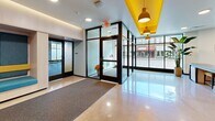 Located at 224 N Barstow St in Eau Claire, Wisconsin, this well-appointed residential lobby serves as a welcoming gateway for its building's residents. The single-floor hallway is thoughtfully designed with a bold, modern aesthetic, highlighted by striking yellow ceiling beams and pendant lights that contrast vividly against the white ceiling and polished concrete floors.

Upon entry, residents and guests are greeted by a set of sleek, floor-to-ceiling glass doors framed in dark metal, flanked by expansive windows that flood the space with natural light and offer views of the surrounding streetscape. A textured entry mat transitions seamlessly to the gleaming lobby floor.

A teal reception desk anchors the interior, complete with a digital display screen showing a welcoming message. Adjacent to the desk, a built-in seating nook features cushioned teal and yellow upholstered benches, offering a comfortable waiting area. A dedicated mail center lines one wall, with rows of individual mailboxes set against a teal base, accompanied by a dark countertop bar with amber upholstered stools—ideal for sorting mail or a quick pause.

Artwork and a framed map add visual interest to the gray accent walls, while recessed and ambient lighting maintain a warm, inviting atmosphere throughout. The overall design is energetic yet functional, making a strong first impression.