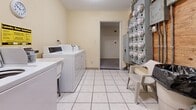 Located at 1447 Woodview Drive in Schererville, Indiana, this shared laundry facility provides a practical and functional space for residents. The room features light-toned walls and clean white tile flooring throughout, contributing to a bright and utilitarian atmosphere.

The laundry area is equipped with multiple top-loading washing machines and dryers arranged along the walls, offering ample capacity for shared use. A folding counter is conveniently positioned near the machines, and posted laundry guidelines help users observe proper etiquette and hours of operation. A wall clock is mounted above the machines for easy time reference.

A large bank of electric utility meters is prominently installed along one wall, indicating that this room serves as a central utility hub for the building. Exposed copper piping runs along the adjacent wall, connecting to the building's plumbing infrastructure. A water heater with associated pipe fittings is also present, housed behind a plywood utility panel in a dedicated corner of the room.

A doorway leads to an adjacent corridor or utility area, providing access through the space. The room's compact but efficiently organized layout ensures that all essential laundry and utility functions are accessible in one centralized location, making it a key shared resource for residents of the building.