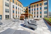 Located in the heart of Missoula, Montana, this generously sized outdoor courtyard terrace is nestled within a modern multi-story building complex, offering a well-appointed communal amenity space that combines functional design with natural beauty. The terrace is defined by a striking patterned deck surface composed of alternating wood and stone tile sections, creating a visually dynamic, checkered layout that spans the full footprint of the courtyard.

At the center of the space stands a warm, golden-toned wood pergola structure, serving as a focal gathering point and providing partial shade over a bar-height seating area. Surrounding the pergola, rows of sleek reclining lounge chairs are thoughtfully arranged to invite relaxation and sun exposure. Additional seating areas, including benches and dining sets, are distributed throughout the terrace, accommodating a variety of social and leisure activities.

Integrated raised planters throughout the space add greenery and seasonal color, softening the architectural elements of the surrounding multi-story façades, which feature a contemporary mix of stucco, stone cladding, wood paneling, and large windows. A perimeter railing along one edge of the terrace opens to sweeping views of Missoula's iconic mountain ridgelines under expansive Montana skies, making this outdoor space as scenic as it is functional.