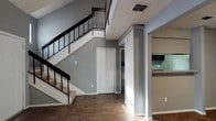 This two-story residence offers a well-organized layout across two levels, making efficient use of its space through thoughtful design. The main level greets you with a front entry area featuring vaulted ceilings that rise dramatically overhead, allowing the living area to feel open and airy. Dark hardwood-style flooring runs throughout the main level, providing a warm, grounded contrast to the cool gray walls.

The living area is generously proportioned for its footprint, benefiting from natural light through a window near the entry. An upper-level railing overlooks the main living space below, adding visual depth and a loft-like quality to the interior. A carpeted staircase with a sleek black metal railing leads to the upper floor, serving as a functional architectural feature.

The kitchen is accessed via a pass-through opening with a granite-style countertop bar, offering a connection between the kitchen and living areas. Inside, the kitchen is equipped with dark cabinetry, granite-patterned countertops, a white refrigerator, an electric range, and a dishwasher, all set against continued hardwood-style flooring. Closet storage is also available on the main level. Textured ceilings are consistent throughout the home.
