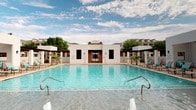 Located at 3850 Sycamore School Rd in Fort Worth, Texas, this single-story outdoor amenity space is designed with a modern Southwest aesthetic, characterized by crisp white stucco walls, clean geometric lines, and warm brick accents. The centerpiece is a generously sized swimming pool with crystal-clear turquoise water, framed by a broad stone tile deck outfitted with teal-cushioned lounge chairs and chaise lounges positioned to take full advantage of the open Texas sky.

Flanking the pool are several covered pergola structures supported by substantial columns, offering shaded retreat areas furnished with teal seating arrangements and low coffee tables. Large louvered windows allow natural light to filter through while maintaining a sense of enclosure and comfort within these shaded spaces.

A fully equipped outdoor kitchen serves as a social hub, featuring stainless steel grills, cabinetry, and an expansive granite-topped bar counter that overlooks the pool area—ideal for entertaining. Palm trees and native stone landscaping accent the perimeter, reinforcing the resort atmosphere. Decorative water jets emerge from the pool's shallow end, adding a dynamic visual element. The overall ambiance is relaxed, open, and suited for outdoor recreation and social gatherings.