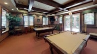 This well-appointed billiards room is designed for both leisure and socializing, centered around two full-size pool tables positioned to allow comfortable play from all sides. The room's most striking architectural feature is its coffered ceiling, detailed with rich dark wood beams that lend a classic, upscale character to the space. Warm-toned carpeting complements the wood accents throughout, creating a cohesive and inviting atmosphere. Along one wall, a built-in upholstered bench with green striped fabric provides comfortable spectator seating, accompanied by decorative display shelving above, adorned with plants, framed artwork, and various decorative objects. High-top tables and bar-style chairs are arranged along the perimeter, offering additional casual seating for guests. Multiple windows dressed with horizontal blinds allow natural light to filter in while maintaining privacy. A wall-mounted flat-screen television adds a modern entertainment element to the room. Glass-panel French doors connect the billiards room to an adjacent space, as well as provide access to an outdoor area, enhancing the sense of openness. Lush indoor plants are thoughtfully placed throughout, adding a refreshing natural element to the refined interior.