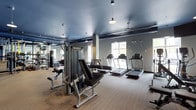 This fitness center offers a thoughtfully designed workout environment that balances functionality with contemporary aesthetics. The space features a cool grey ceiling accented by pendant lighting and ceiling fans, contributing to a sleek, modern atmosphere. Wood-toned flooring runs throughout, providing both durability and visual warmth.

The cardio zone is prominently positioned near large windows that allow natural light to fill the space, with multiple treadmills aligned to take advantage of the outdoor views. A dedicated strength training area includes a multi-station cable and weight machine, along with additional resistance equipment designed to accommodate a range of exercise routines.

A floor-to-ceiling mirrored wall along one side visually expands the space while allowing users to monitor their form during workouts. A punching bag is also visible in the mirrored section, adding a versatile training option. Structural columns are integrated naturally into the layout without disrupting the flow of the space. The overall ambiance is clean, functional, and inviting, making it well-suited for a variety of fitness activities.