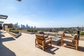 Located at 1011 Everett St in the vibrant Echo Park neighborhood of Los Angeles, this contemporary amenity space combines a well-appointed interior living room with a spectacular rooftop terrace. The interior living room features light hardwood flooring, crisp white walls, and a large window fitted with sleek horizontal blinds that flood the space with natural light. The room is furnished with a modern gray sofa, bold orange accent chairs, a glass coffee table, and a wall-mounted flat-screen TV, creating a comfortable and stylish lounge environment. Art accents the walls, adding a creative touch to the clean, minimalist aesthetic. The rooftop terrace is the true highlight of the space, offering sweeping panoramic views of the Downtown Los Angeles skyline, the surrounding hillside neighborhoods, and mountain ranges in the distance. The terrace is thoughtfully designed with multiple seating areas, including teak lounge chairs and a full outdoor dining set with umbrella shading. Built-in planters with native landscaping add greenery and texture, while a fire pit area provides an intimate gathering spot. The contemporary architectural design incorporates warm terracotta-toned accent walls and smooth stucco finishes throughout the outdoor space.