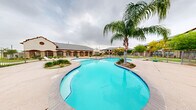 This outdoor pool area serves as a central amenity within a well-maintained residential community, offering a thoughtfully designed space for recreation and relaxation. The centerpiece is a generously sized freeform swimming pool with smooth, curving edges and vivid turquoise water, lending the space a resort-like character. A distinctive design feature is the landscaped island within the pool itself, anchored by a tall, mature queen palm that provides a striking focal point and natural shade. Adjacent to the main pool is a separate, shallower wading pool, ideal for younger users, connected by a broad concrete sundeck that provides ample space for lounge chairs and casual gatherings. The pool area is fully enclosed by a metal security fence, ensuring safety while maintaining open sightlines to the surrounding community grounds. Neatly trimmed ornamental shrubs, decorative planters, and patches of greenery add to the landscaped aesthetic throughout the deck. In the background, the community's multi-story buildings feature a traditional architectural style with stone and brick accents, arched entryways, and pitched rooflines. A covered pavilion structure with decorative arches is also visible, offering shaded seating near the pool. A playground area with colorful equipment is situated just beyond the fencing, rounding out the outdoor amenities available within this vibrant community setting.