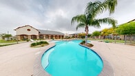 This outdoor pool area serves as a central amenity within a well-maintained residential community, offering a thoughtfully designed space for recreation and relaxation. The centerpiece is a generously sized freeform swimming pool with smooth, curving edges and vivid turquoise water, lending the space a resort-like character. A distinctive design feature is the landscaped island within the pool itself, anchored by a tall, mature queen palm that provides a striking focal point and natural shade. Adjacent to the main pool is a separate, shallower wading pool, ideal for younger users, connected by a broad concrete sundeck that provides ample space for lounge chairs and casual gatherings. The pool area is fully enclosed by a metal security fence, ensuring safety while maintaining open sightlines to the surrounding community grounds. Neatly trimmed ornamental shrubs, decorative planters, and patches of greenery add to the landscaped aesthetic throughout the deck. In the background, the community's multi-story buildings feature a traditional architectural style with stone and brick accents, arched entryways, and pitched rooflines. A covered pavilion structure with decorative arches is also visible, offering shaded seating near the pool. A playground area with colorful equipment is situated just beyond the fencing, rounding out the outdoor amenities available within this vibrant community setting.
