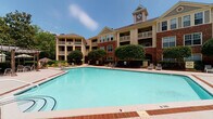 Located at 100 Crescent Arbor Ln in Cary, North Carolina, this outdoor amenity space is the centerpiece of a well-appointed residential community. The focal point is a generously sized swimming pool with clear, blue water, framed by a brick-accented concrete pool deck designed for both relaxation and socializing. Lounge chairs are arranged throughout the deck, offering ample space for sunbathing and leisure under the open sky.

A shaded pergola area anchored by wooden posts provides a comfortable retreat from the sun, furnished with outdoor tables, chairs, and large patio umbrellas. This covered seating zone overlooks the pool on one side and backs up to mature, lush greenery on the other, creating a serene and private atmosphere. Tall trees and manicured hedges border the space, enhancing the sense of natural seclusion.

The surrounding multi-story brick buildings feature classic architectural detailing, including balconies, decorative dormers, and a distinctive clock tower that serves as a charming landmark for the community. Neatly trimmed ornamental shrubs and landscaped beds line the buildings, adding to the polished, well-maintained aesthetic. The overall ambiance is one of comfortable, resort-style outdoor living in a lush, tree-lined setting in the heart of Cary, NC.